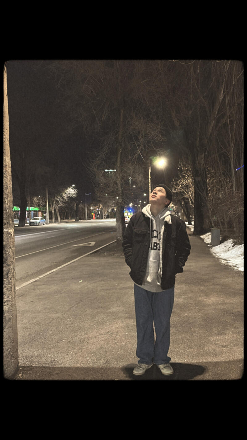 A person stands on a sidewalk at night, looking up at a streetlight, wearing a beanie, a hoodie, a jacket, and jeans.