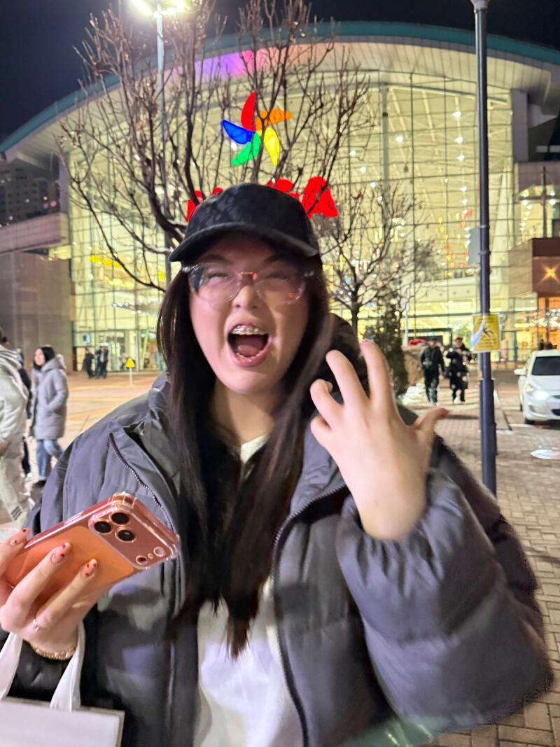 A young woman with glasses and braces, wearing a black cap and gray puffer jacket, is holding a phone with a clear case and smiling with her mouth open; in the background is a modern building with a colorful windmill decoration.