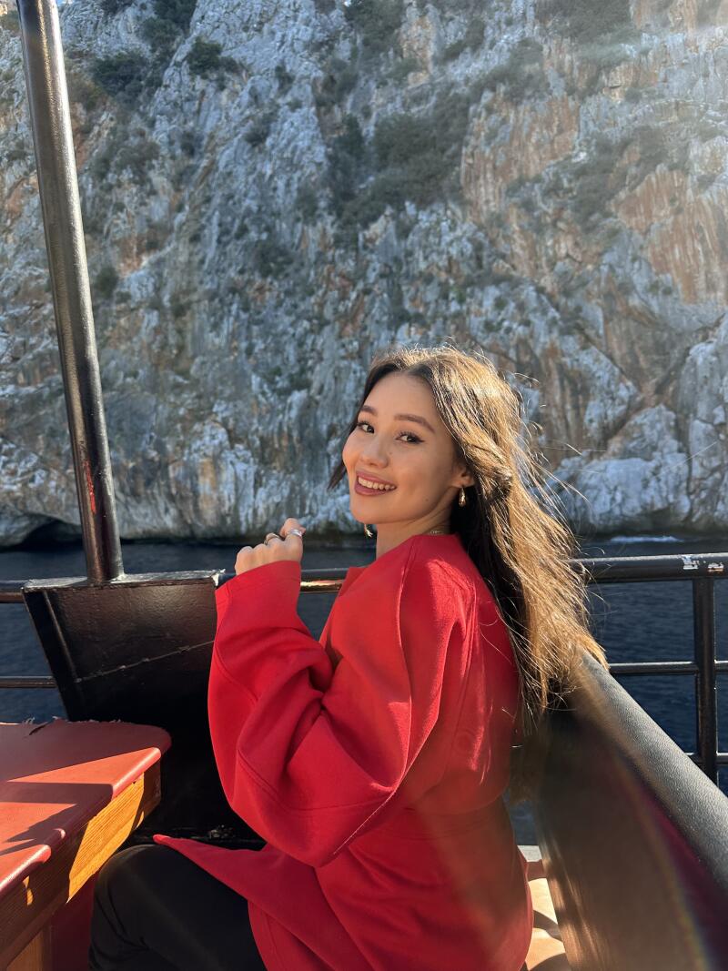 A smiling woman in a red coat sits on a boat, with a rocky cliff and water in the background.