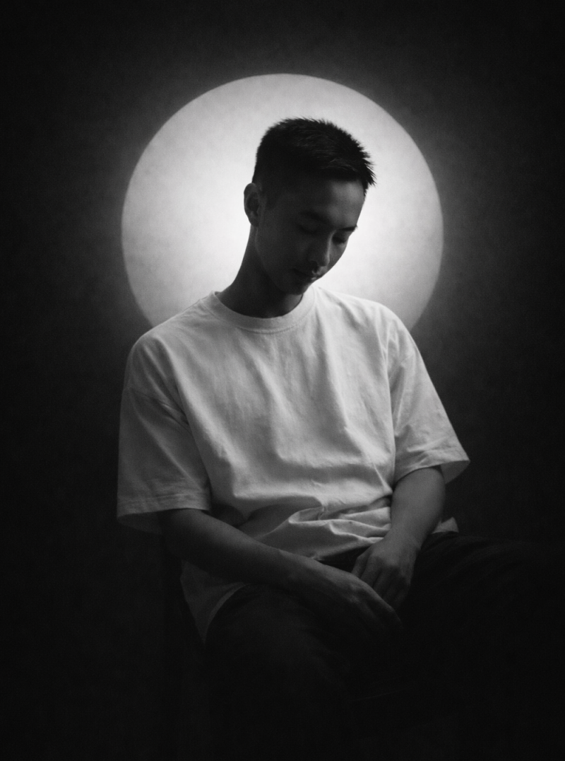 A grayscale portrait of a person with short hair wearing a white t-shirt, looking down, against a dark backdrop with a bright, circular light behind them.