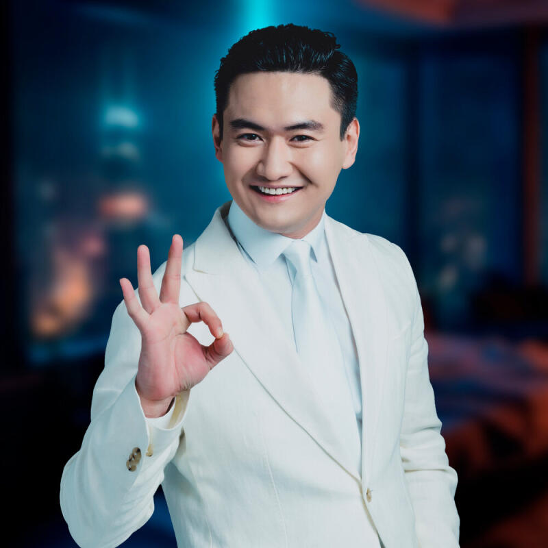 A smiling man in a white suit and tie is making an "okay" hand gesture.