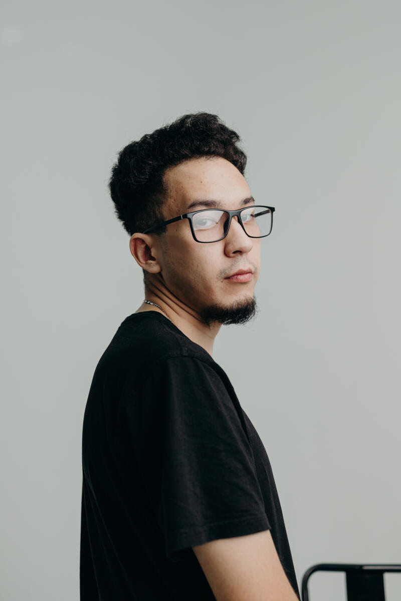 A man with curly hair and glasses, wearing a black T-shirt, looks over his shoulder.