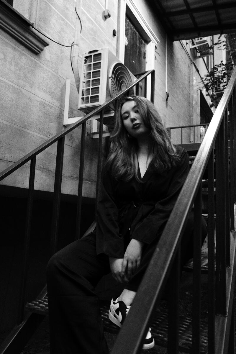 A monochrome medium shot depicts a woman with long hair sitting on metal stairs, dressed in a dark jacket and pants, with white shoes with black details; an air conditioner is visible on the wall in the background.