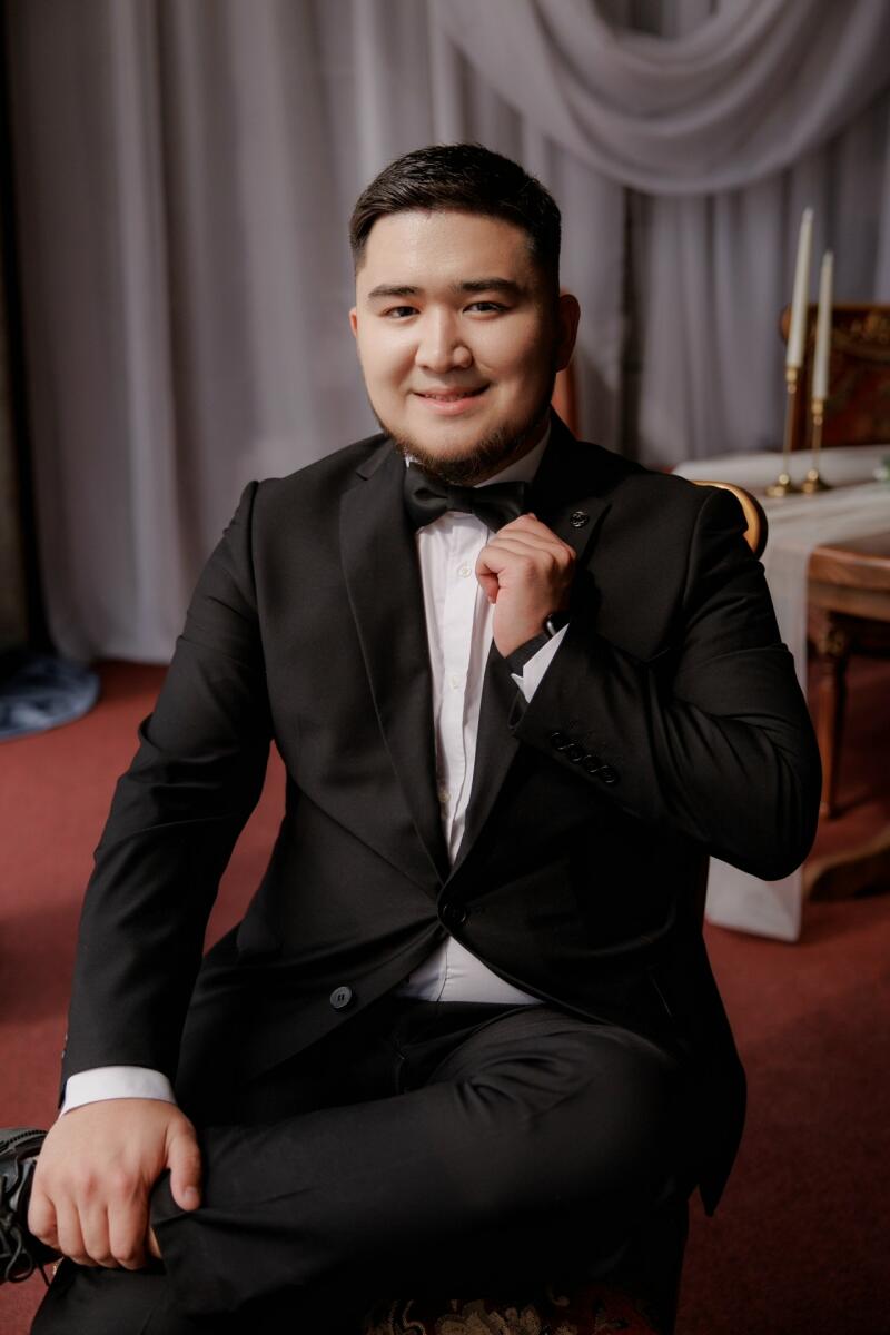 A man in a black suit and bow tie is sitting down, smiling slightly and holding his bow tie.