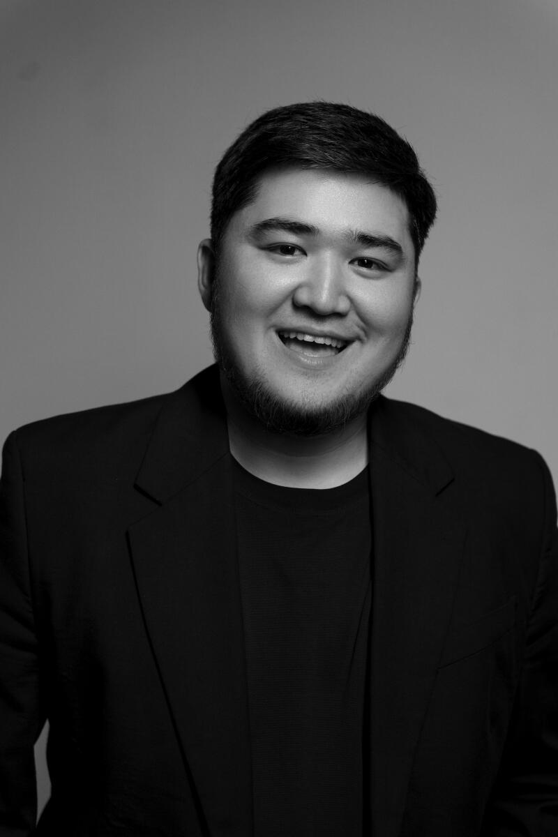 A black and white portrait of a smiling man with a beard, wearing a black t-shirt and blazer.