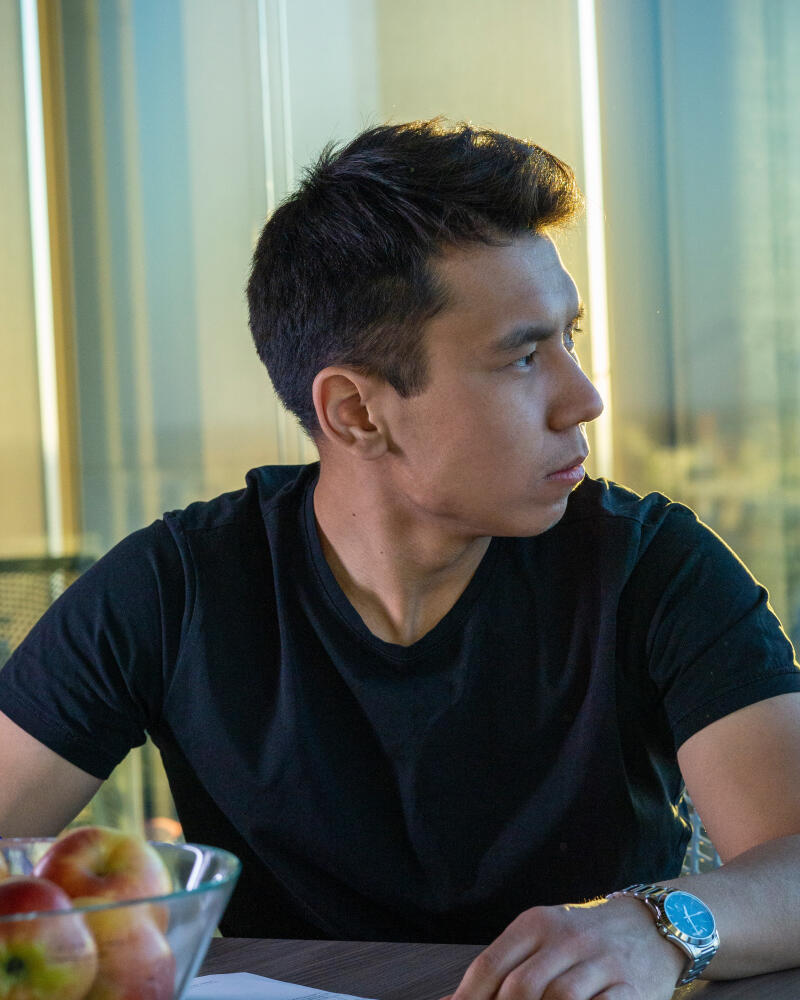 A man in a black t-shirt and a wristwatch is sitting at a table with a bowl of apples.