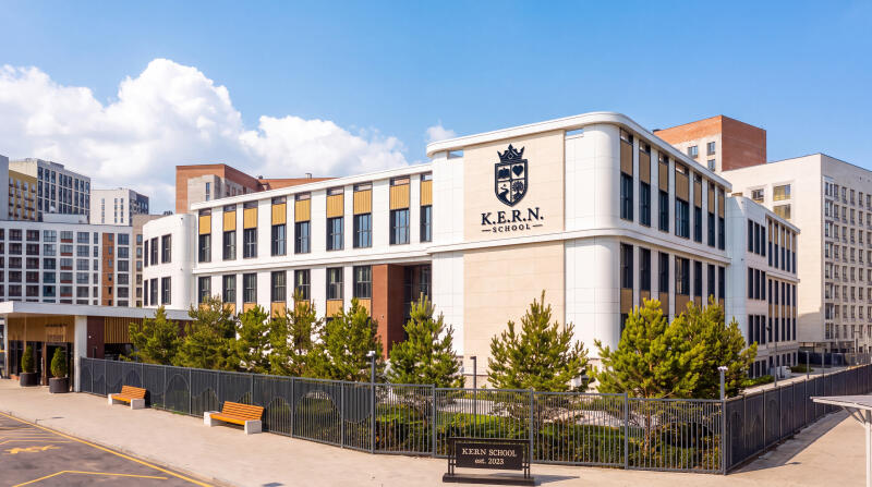 A modern K.E.R.N. School building with white and beige facades stands under a clear blue sky, surrounded by green trees, a fence, and two wooden benches, with a sign reading "KERN SCHOOL est. 2023" in the foreground.