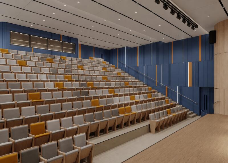 The image shows the interior of a lecture hall with rows of gray and yellow seats arranged on risers, facing a wooden stage. The walls are decorated with vertical blue panels accented by white and orange strips, and overhead lighting illuminates the space.