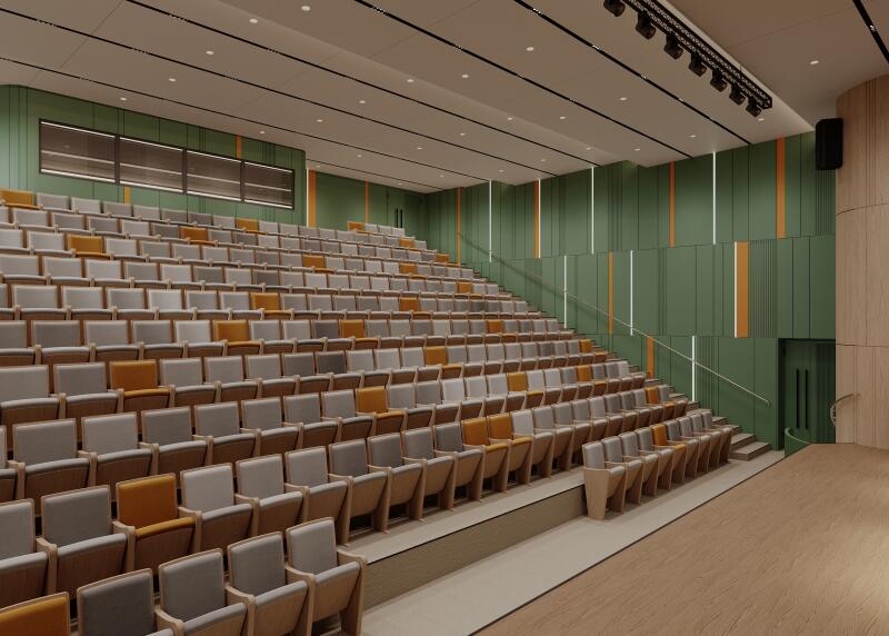The photo shows a large auditorium with rows of gray and orange seats ascending towards the back. The walls are green with vertical stripes of orange and white, and the ceiling has track lighting.