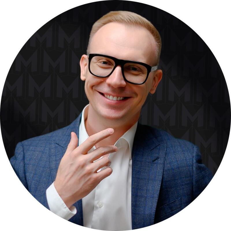 A man with short blond hair and glasses smiles, wearing a blue blazer and white shirt, set against a dark background with a repeating 'M' pattern.