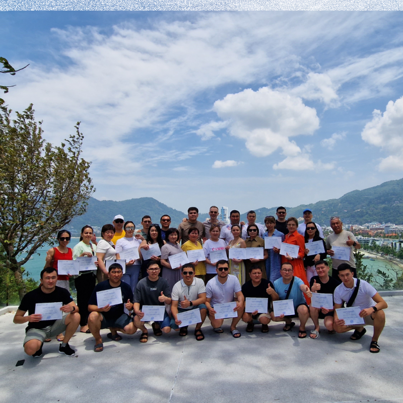 A group of about 30 people, mostly wearing casual clothing, are posing outdoors, holding certificates and giving thumbs up against a backdrop of mountains and a beach.
