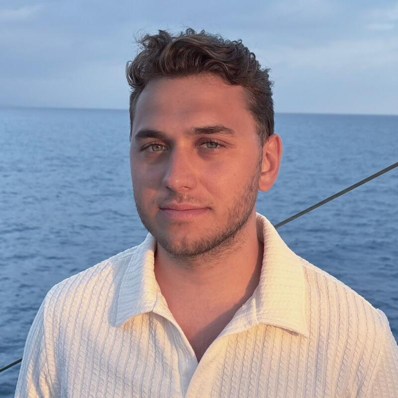 A man with curly hair and a white button-down shirt stands in front of a body of water.
