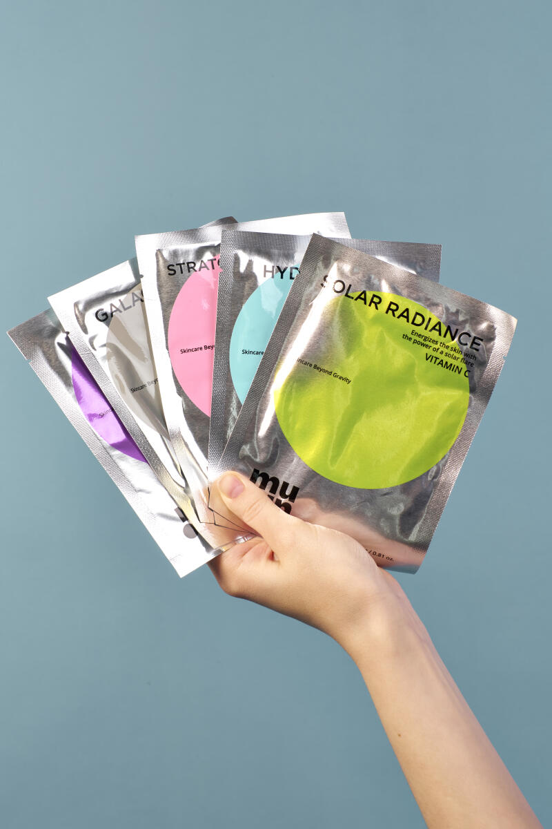 A hand holds several foil packets of Mu skincare products, including varieties labeled 'GALA,' 'STRAT,' 'HYD,' and 'SOLAR RADIANCE,' against a light blue background.