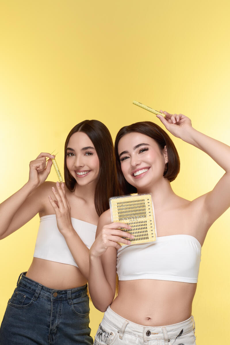 Two smiling women in white tube tops and jeans pose against a yellow background, holding false eyelashes and an applicator.