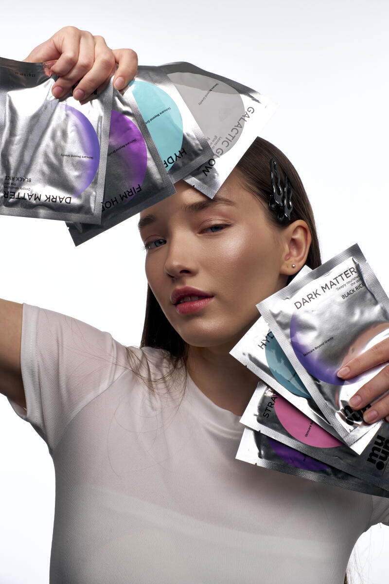 A woman in a white t-shirt is holding up multiple skincare masks with labels like "DARK MATTER BLACK RICE", "FIRM HO", "HYDF", and "GALACTIC GLOW". She has dark hair pulled back with a flame-shaped clip.
