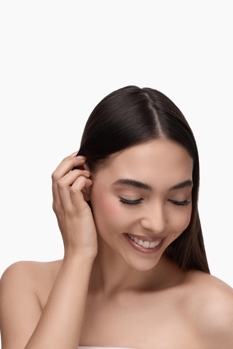 A woman with long brown hair is smiling with her eyes closed, touching her hair with her left hand.