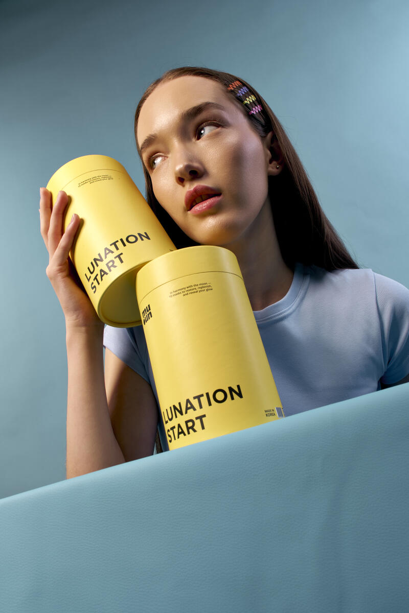 A woman with colorful hair clips holds two yellow containers labeled "LUNATION START" in front of a blue background.