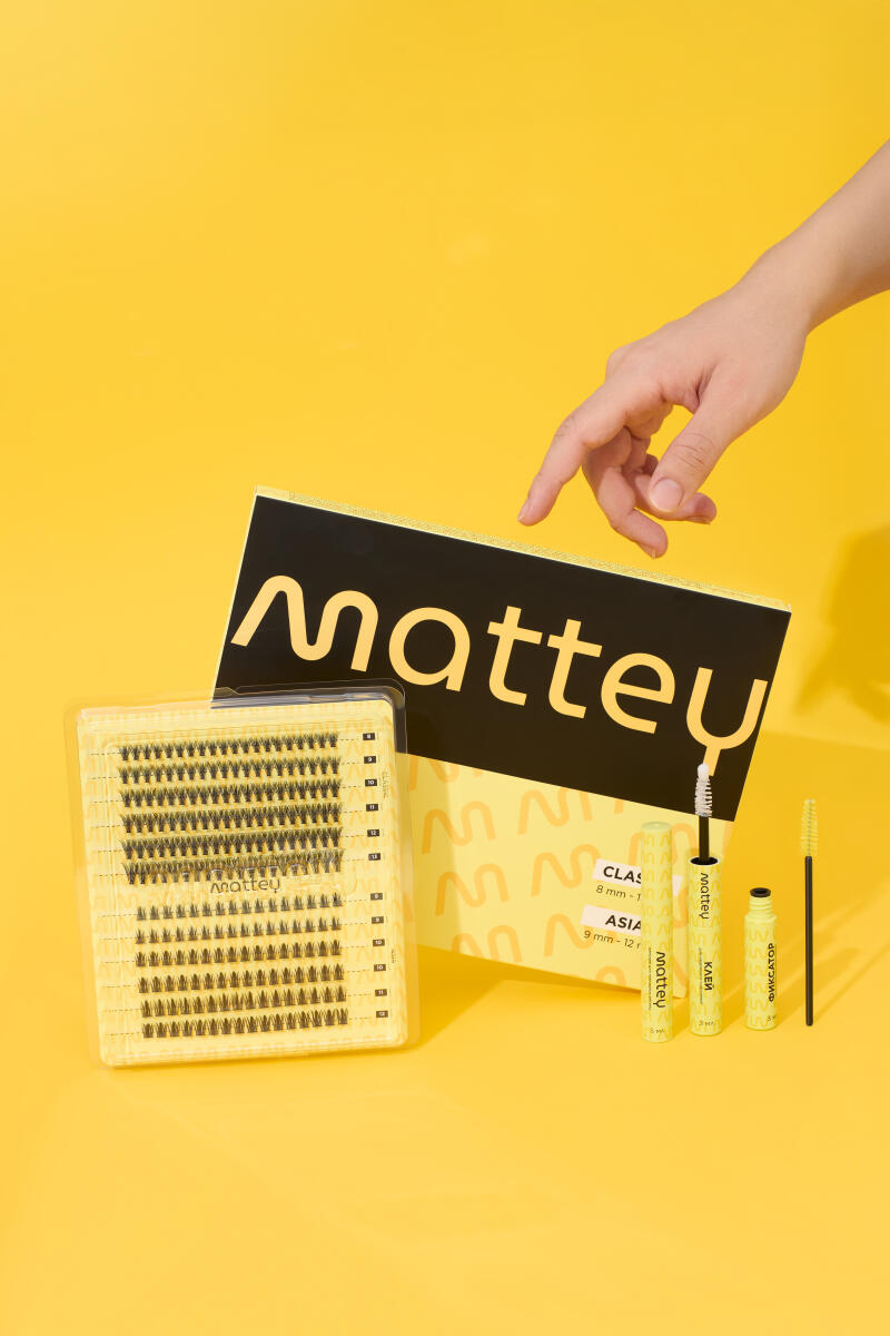 A hand hovers over a package of mattey brand false eyelashes and eyelash accessories on a yellow background; the packaging includes the text 'mattey,' 'КЛЕЙ,' 'CLAS,' 'ASIA,' and 'ФИКСАТОР.'