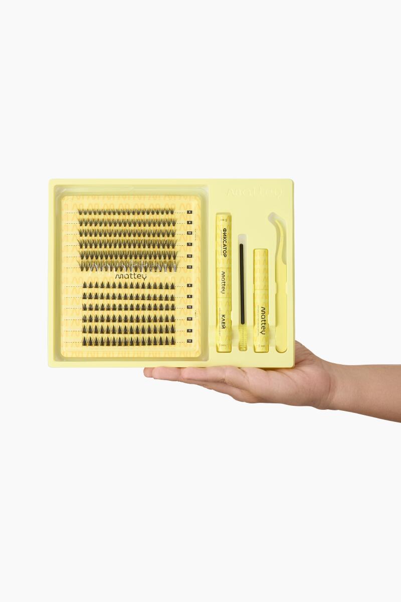 A hand holds a yellow kit containing individual false eyelashes, tweezers, and adhesive, labeled with the brand name 'Mattey' and other inscriptions in Cyrillic.