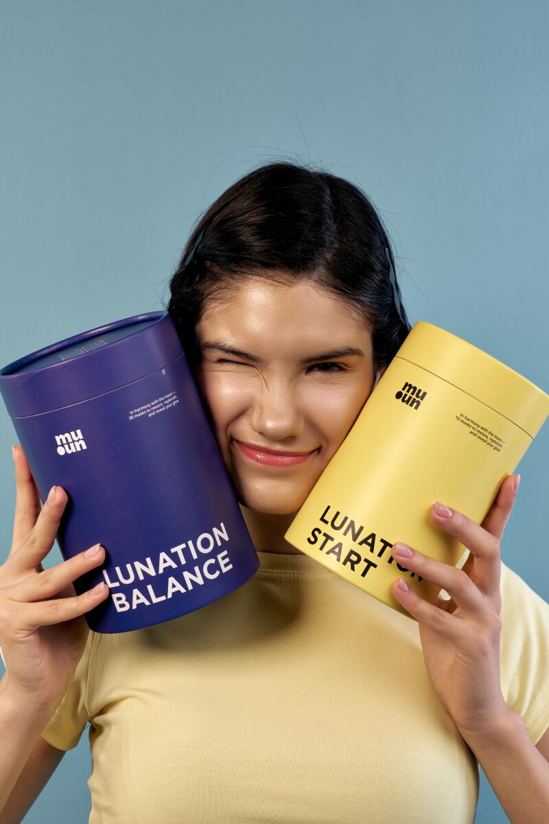 A woman winks while holding two Muoun containers, one blue labeled "Lunation Balance" and the other yellow labeled "Lunation Start," against a blue background.