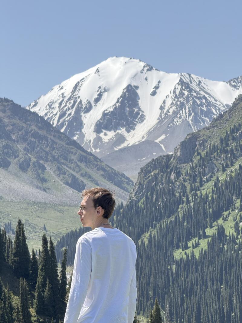 A man in a white shirt stands in a mountainous area with evergreen trees, looking towards a large snow-capped mountain under a clear sky.