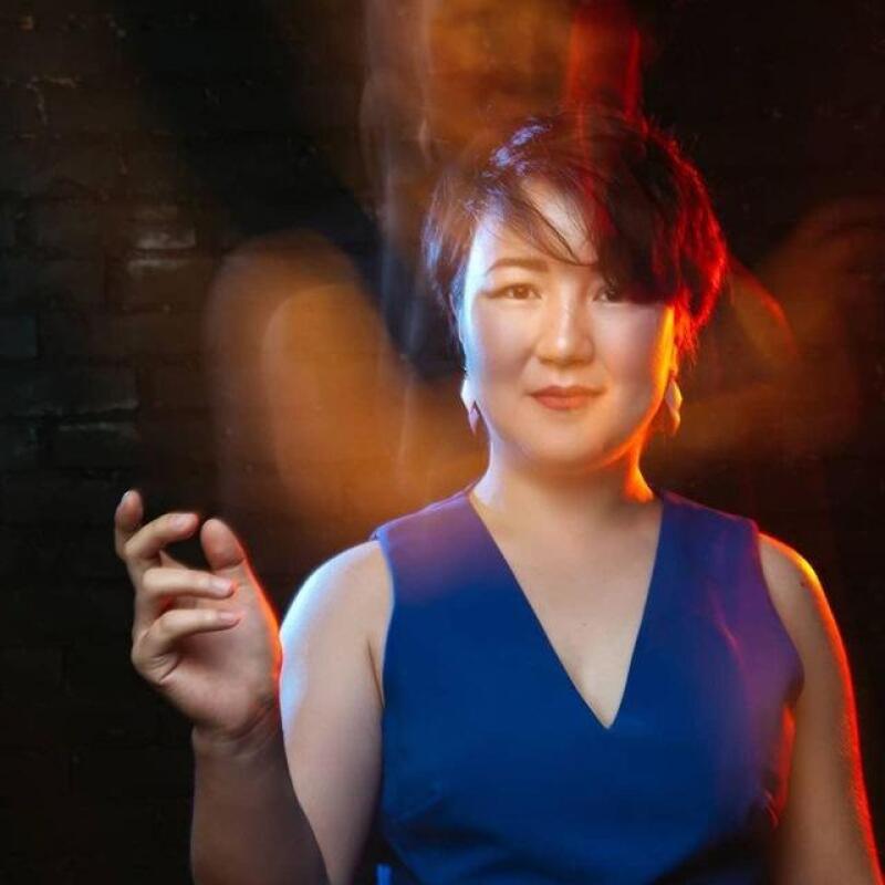 A woman with short dark hair and a blue V-neck top smiles, holding her left hand up against a dark, brick-like background with warm red and orange lighting and motion blur effects.