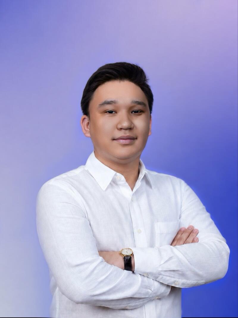 A young man with dark hair, wearing a white shirt and a gold watch, stands with his arms crossed against a blue and purple gradient background, looking at the camera.