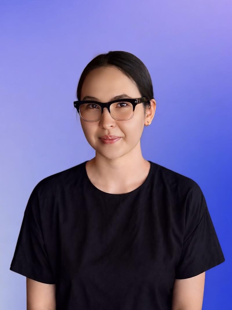 A young woman with dark hair pulled back, wearing black-framed glasses and a black t-shirt, looks forward with a slight smile against a gradient blue and purple background.