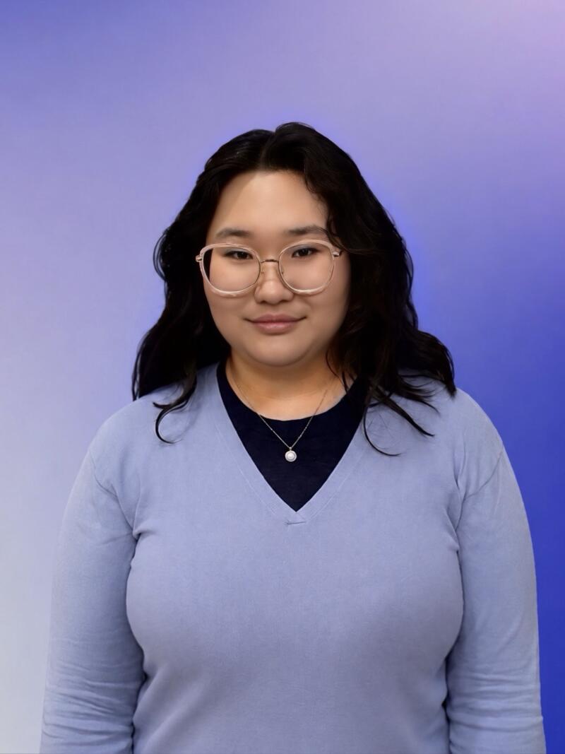 A woman with dark hair and glasses wears a light blue V-neck sweater over a dark shirt, with a pearl necklace, standing against a gradient background of purple and blue.