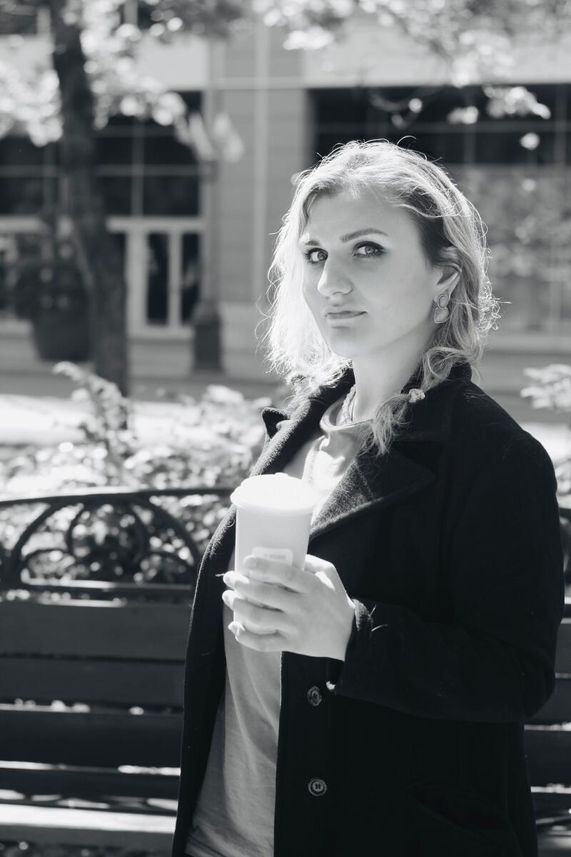 A blonde woman in a dark coat holds a cup and looks at the camera in a black and white outdoor portrait, with a blurred background of a building and trees.