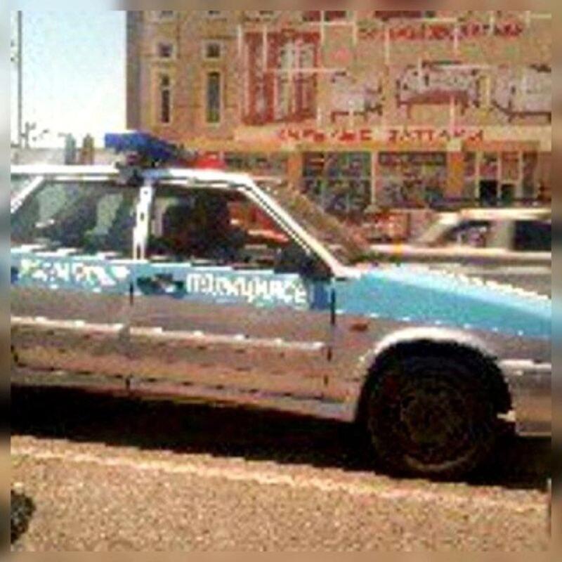 На улице припаркована полицейская машина в сине-белой расцветке, на боку виден текст на кириллице.