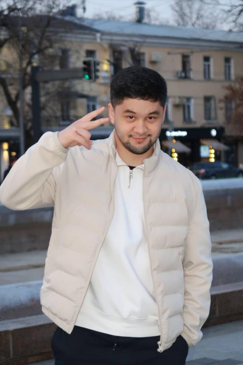 A man in a light jacket and white sweater makes a peace sign in front of a city street with a building and traffic light in the background.