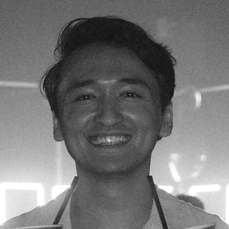 A grayscale portrait of a smiling man with dark hair, wearing a light-colored shirt with dark straps or suspenders.