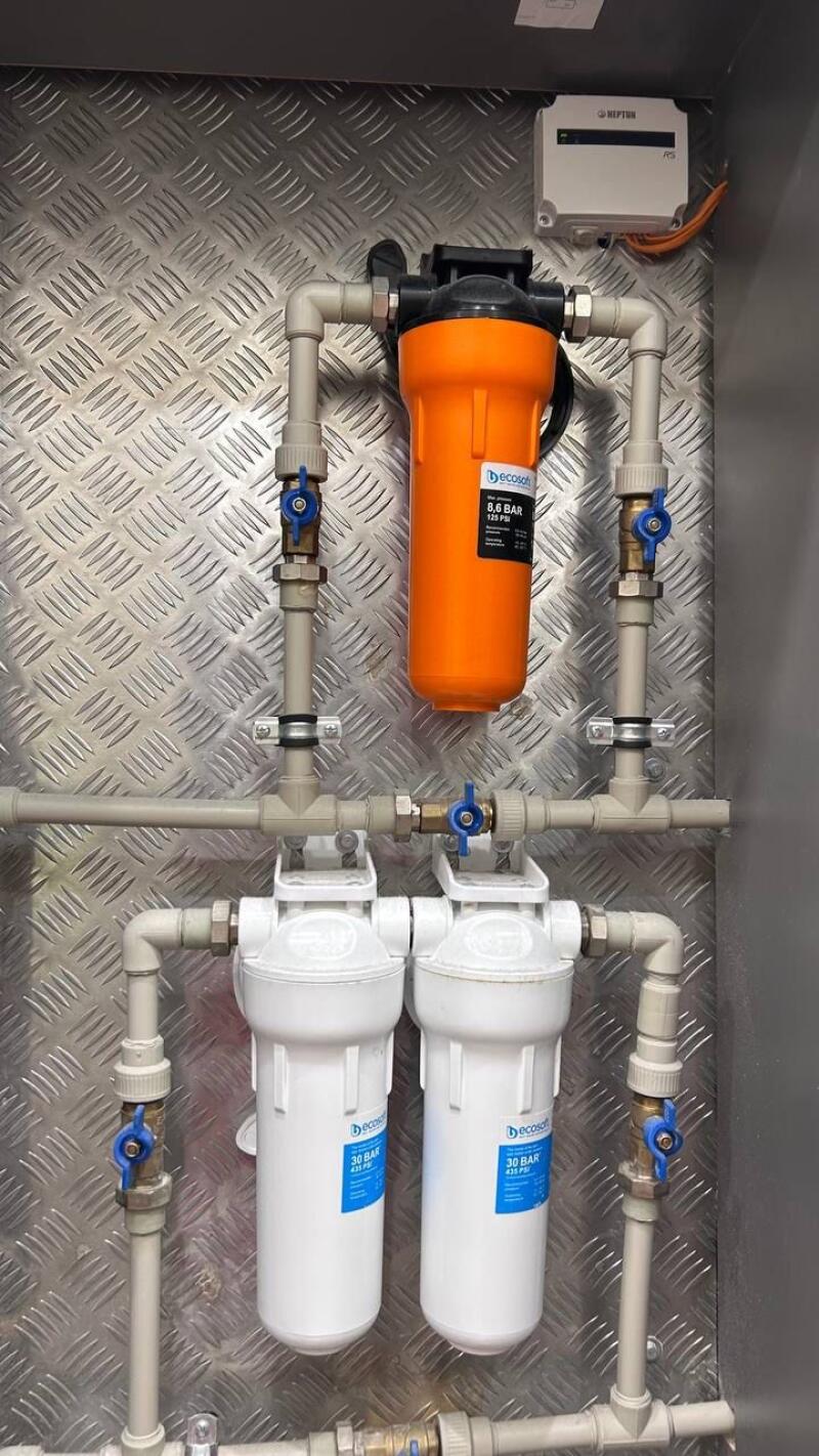 A water filtration system is mounted on a textured metal wall, featuring one orange filter labeled "Decosat 8.6 BAR 125 PSI" and two white filters each labeled "Decosat 30 BAR 435 PSI", connected by light-colored pipes with blue-handled valves. A white control box labeled "NEPTUN RS" is visible in the upper right.
