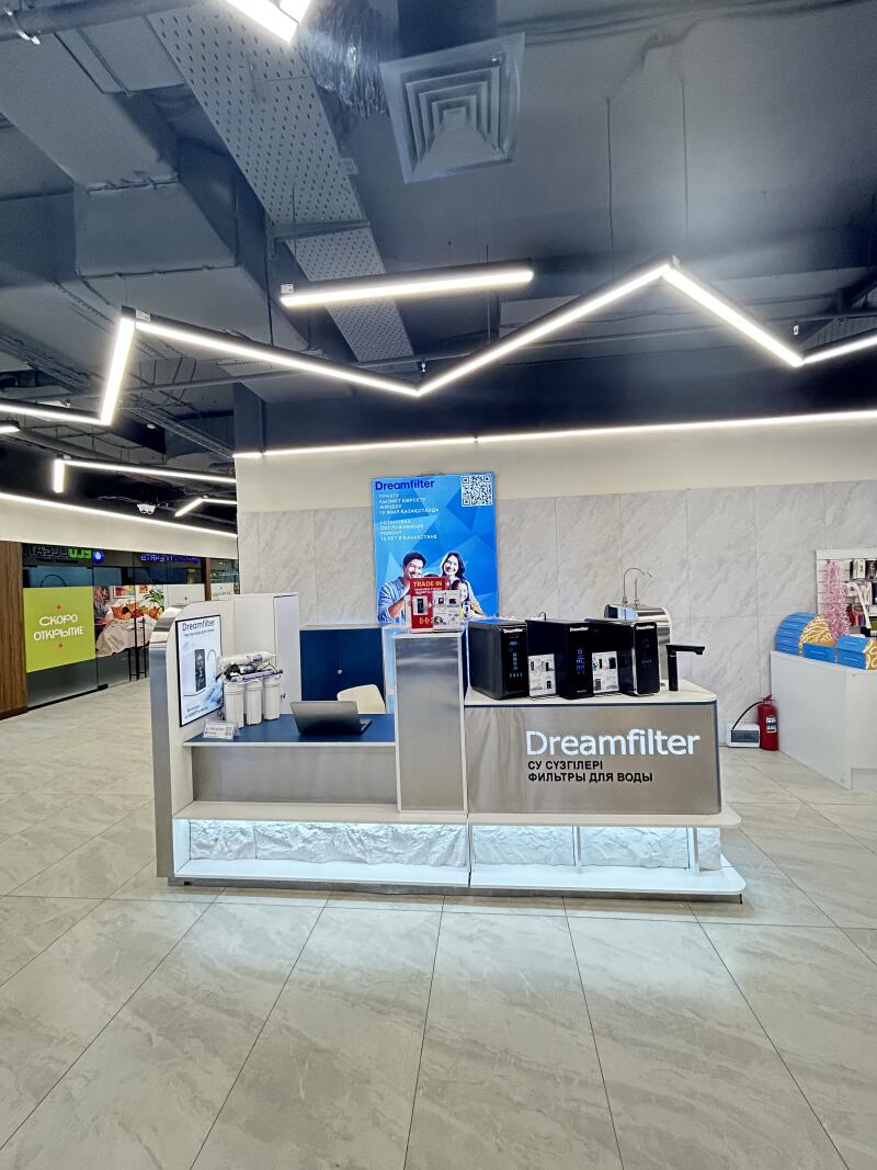 A retail display for Dreamfilter water filters features various filter units, a laptop, and an advertising poster with the brand logo, a QR code, and text "TRADE IN". The main stand also displays "Dreamfilter СУ СҮЗГІЛЕРІ ФИЛЬТРЫ ДЛЯ ВОДЫ". Modern linear lighting illuminates the ceiling.