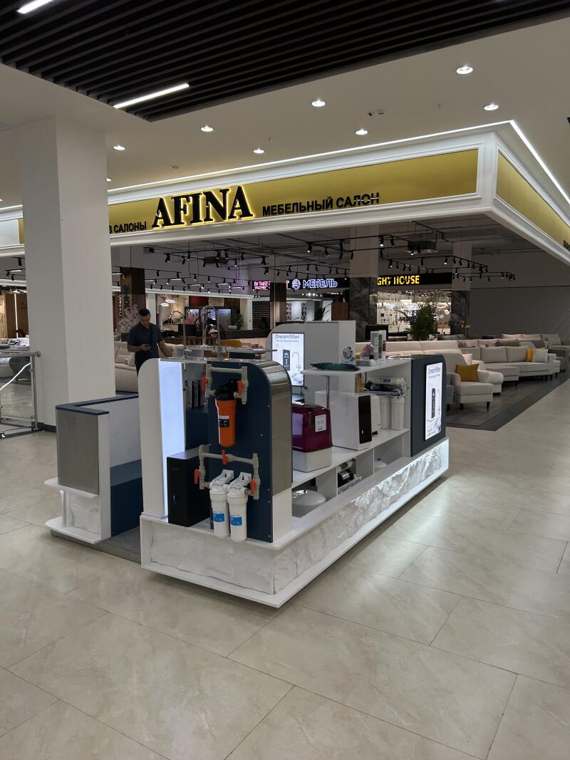 An indoor shot of a furniture store with a prominent yellow sign "AFINA МЕБЕЛЬНЫЙ САЛОН" (AFINA FURNITURE SALON) in the background, and a display stand featuring water filtration systems, including an orange filter, in the foreground.