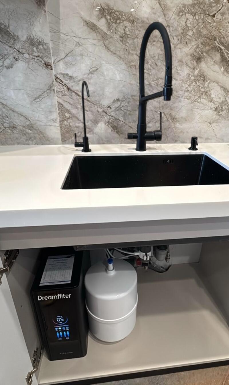 A kitchen sink area featuring a white countertop, a black sink, and three black faucets against a marble-patterned wall. Below the sink, inside an open cabinet, a black "Dreamfilter" water purification system with a digital display showing "05" and a white tank are visible.