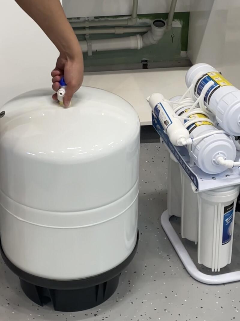 A person's hand is connecting a fitting to the top of a white water pressure tank, next to a multi-stage water filtration system with visible text 'BLUE TIDE RO SYSTEM'.