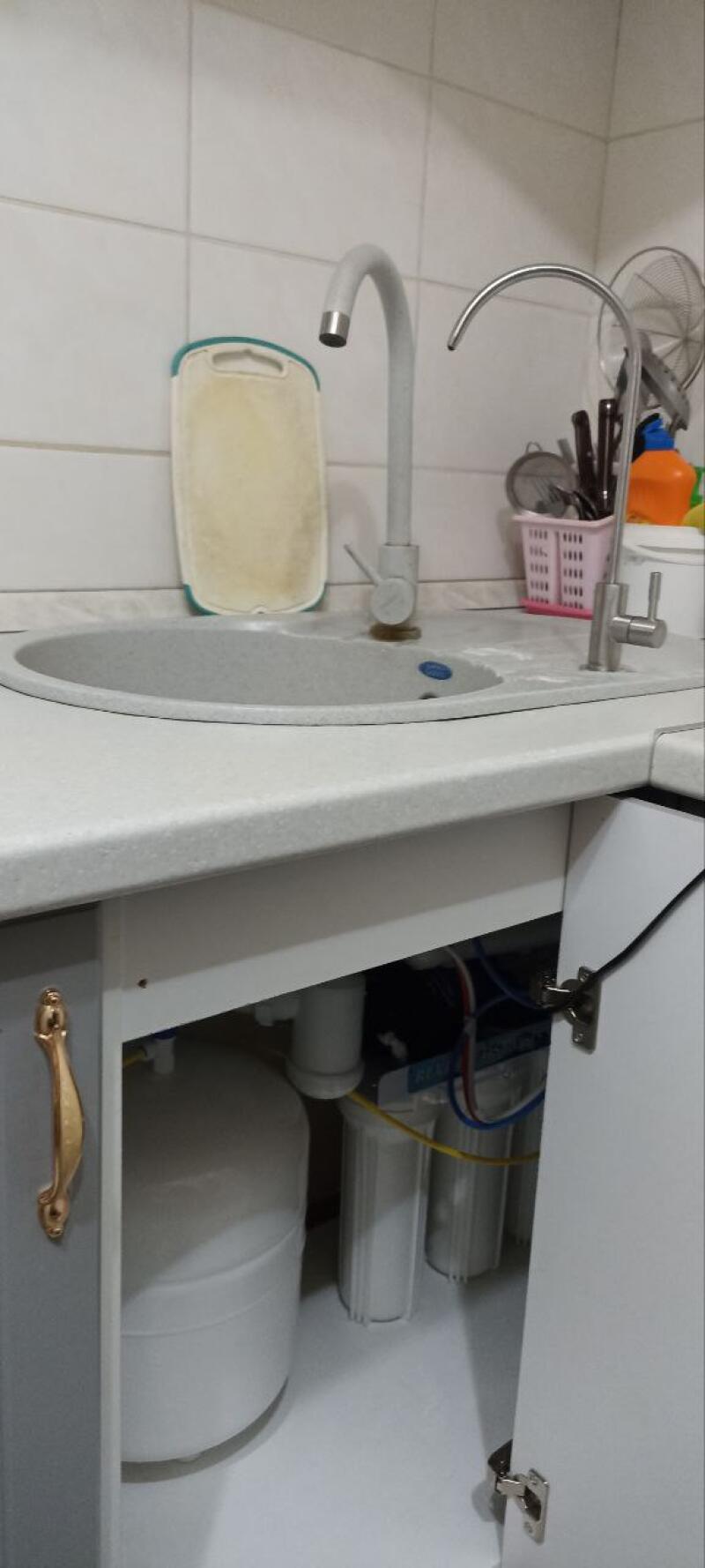 A kitchen scene showing a sink with two faucets, a cutting board leaning against the white tiled wall, and a pink basket of utensils. Below the sink, an open cabinet reveals a white water filtration system with multiple filters and a tank.