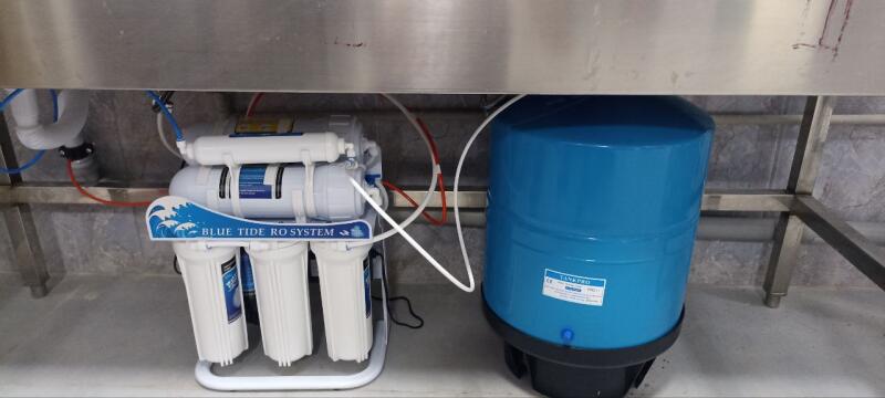 A water filtration unit, labeled "BLUE TIDE RO SYSTEM" with multiple white cartridges, and a blue "TANKPRO" pressure tank are connected by various tubes under a stainless steel surface.
