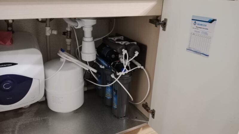 View inside a cabinet under a sink, showing a white Ariston water heater on the left, a white water tank, and a multi-stage water filtration system with several dark blue filter housings and white tubes on the right. Plumbing pipes and a drain are visible, and a sticker with 'DREAMFILTER' and 'Фильтры для воды' is attached to the inside of the cabinet door.