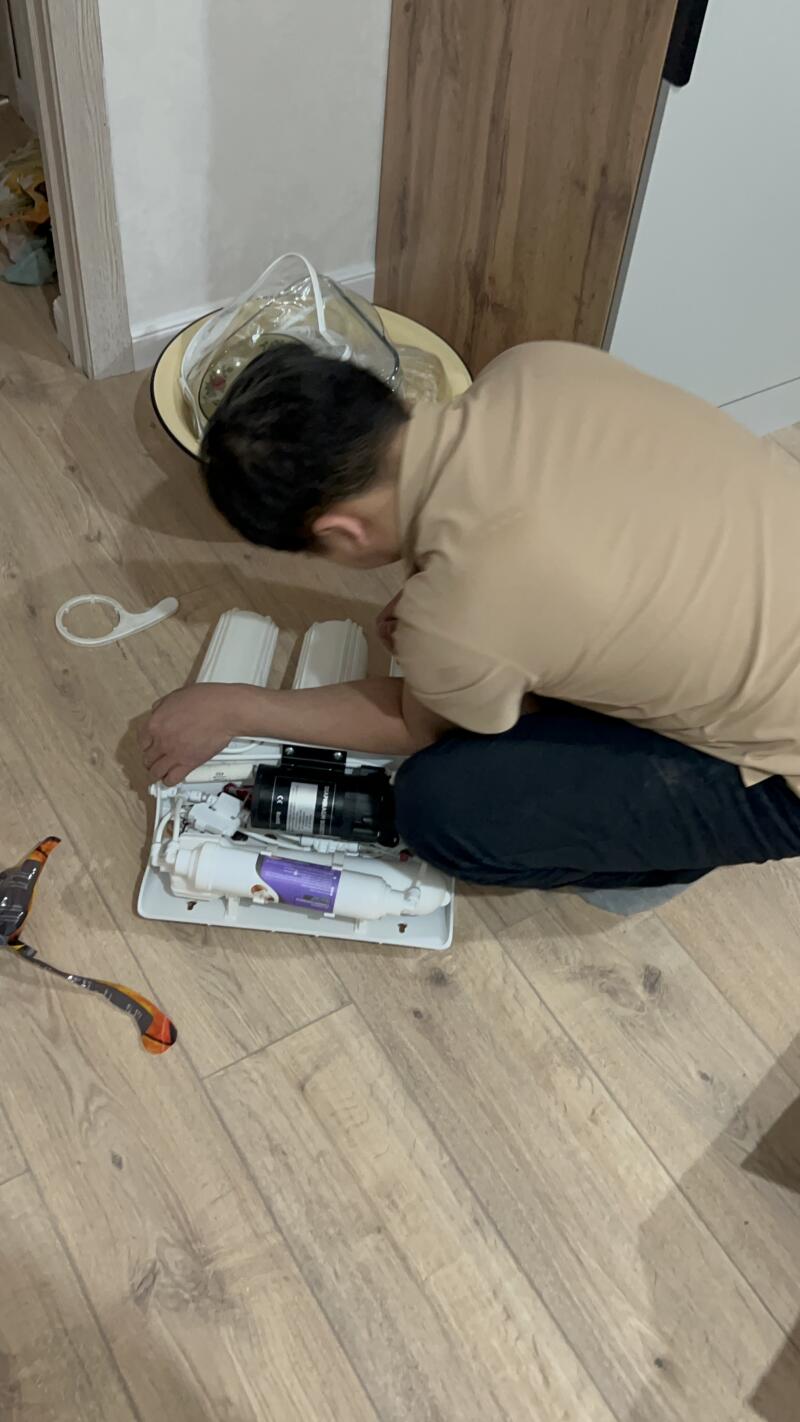 A person in a beige t-shirt and black pants is kneeling on a wooden floor, examining a white water filtration system with multiple filters and a black pump. A white wrench tool is nearby, and a yellow basin is in the background.