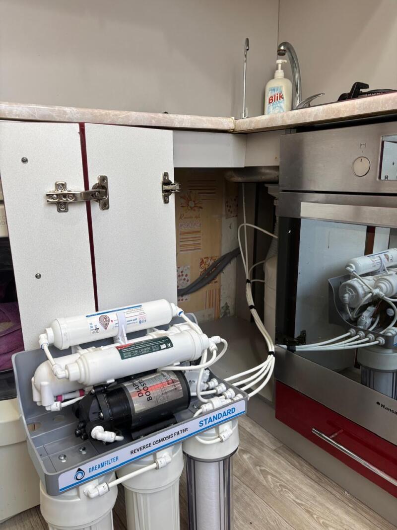 A Dreamfilter "STANDARD" reverse osmosis water filtration system with a "BOOSTER" pump and cartridges is installed on a wooden floor under an open kitchen cabinet. Above, a sink with a faucet and "Blik" soap bottle is on the countertop, next to a partial view of a stainless steel oven.
