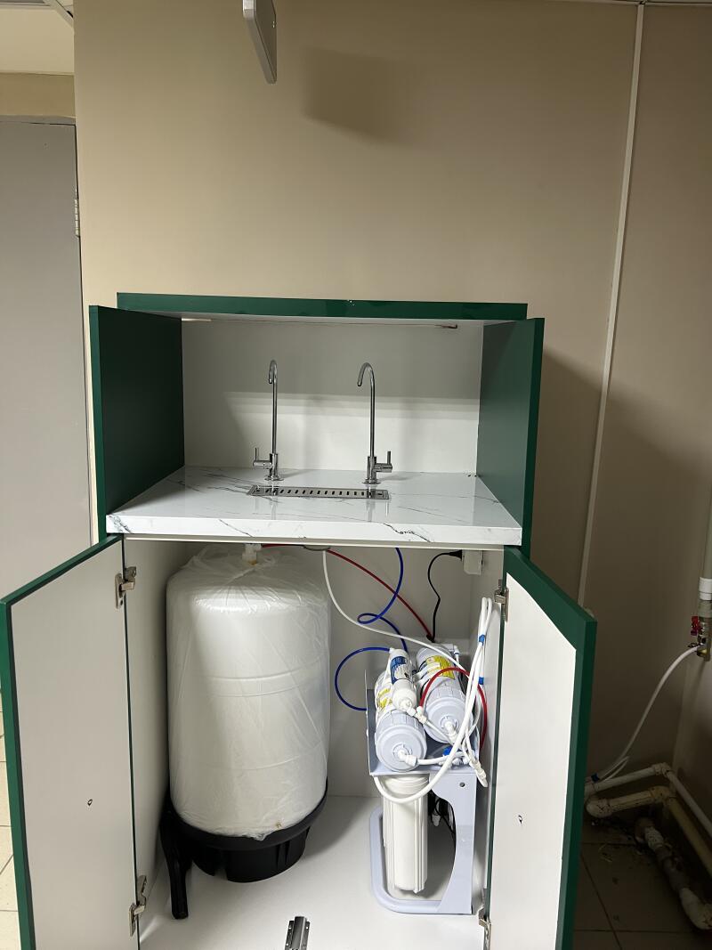 A green and white cabinet features two silver faucets on a marble-patterned countertop. Inside the open cabinet, there is a white water tank and a multi-stage water filtration system with colored tubes.
