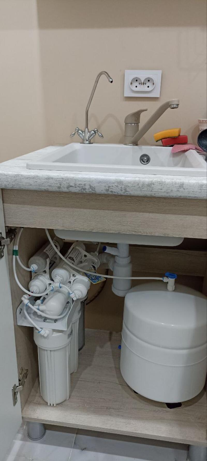 A kitchen sink with two faucets and a power outlet on the wall above it. Below the sink, inside an open cabinet, a multi-stage water filtration system with white cartridges and a large white storage tank is visible.