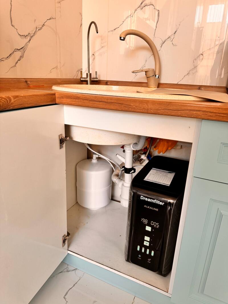 An open kitchen cabinet reveals a water filtration system under a sink, consisting of a white tank and a black unit labeled "Dreamfilter ALKALINE" with a digital display showing "198 005".