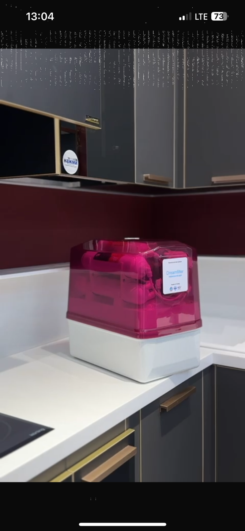 A pink and white "Dreamfilter" water filter with a clear top, labeled "Made in Turkey," sits on a white kitchen countertop. Dark grey cabinets with gold handles and a dark red backsplash are in the background, with a white circular sticker reading "нажми" on an upper cabinet.