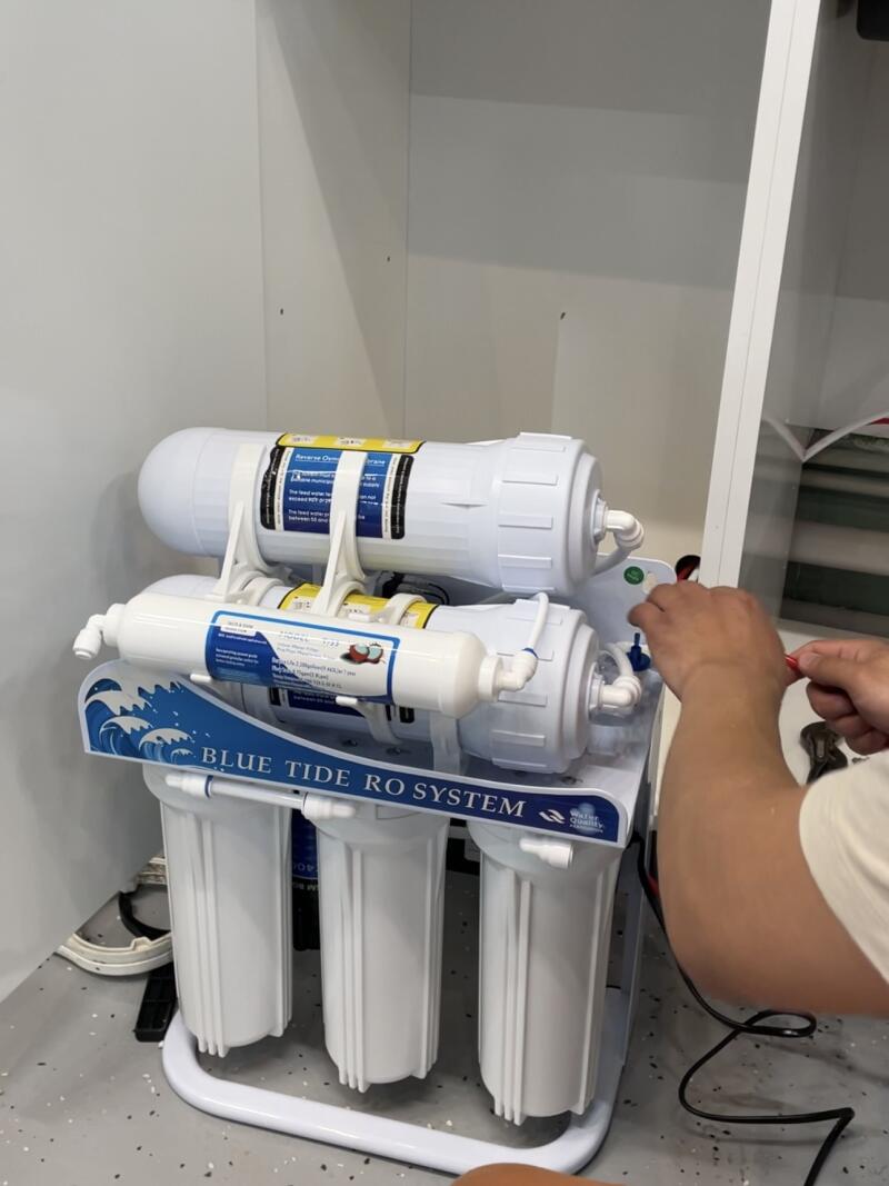 A "BLUE TIDE RO SYSTEM" water filter unit with multiple white cartridges is being worked on by hands on the right, set against a white wall and grey speckled floor.