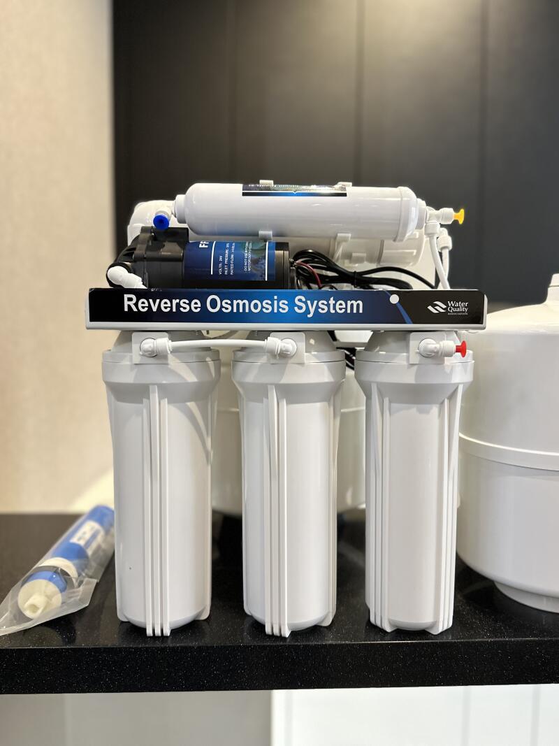 A white reverse osmosis water filtration system with three large filter housings is displayed on a dark surface, featuring a blue label that reads "Reverse Osmosis System" and "Water Quality." A separate blue filter is visible in a plastic bag on the left.