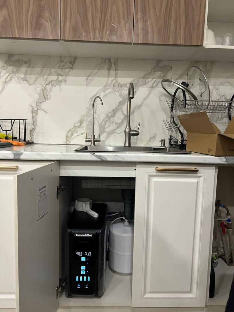 A kitchen sink area with two faucets, a marble-patterned backsplash, and wooden overhead cabinets. An open cabinet below the sink reveals a black "Dreamfilter ALKALINE" water filter system with a digital display showing "483 013" and a white pressure tank.